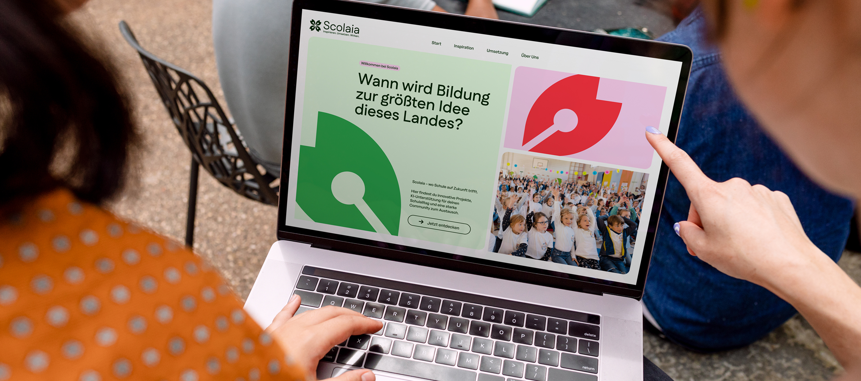 Two people interacting with a laptop displaying the Scolaia website homepage in German.