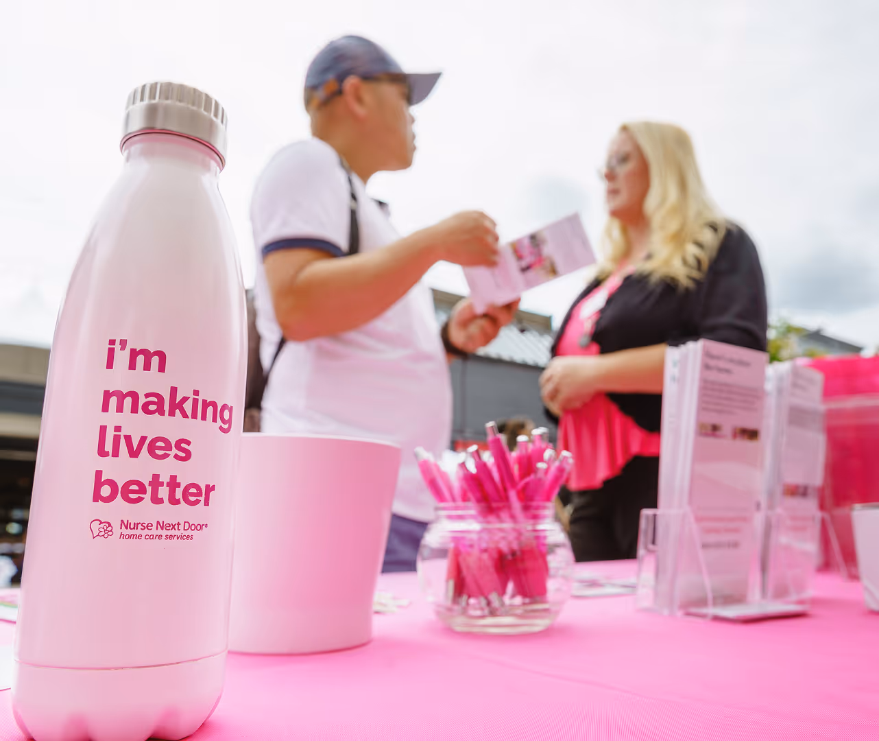 Nurse Next Door representative speaking with a visitor at a pink booth featuring branded materials and a water bottle that reads ‘I’m making lives better.