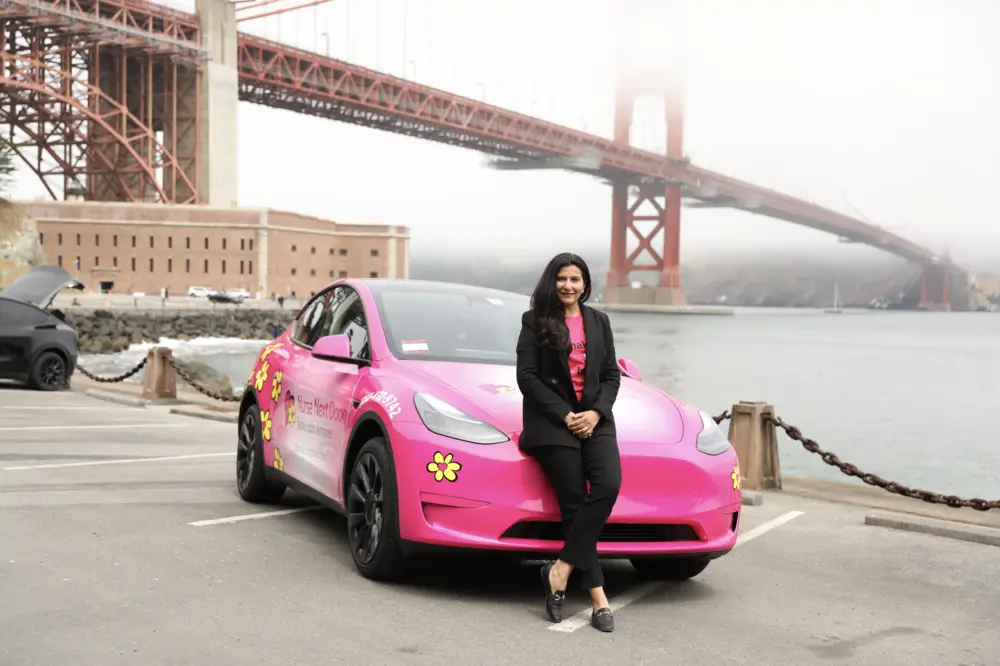 Person in bright pink Nurse Next Door uniform sitting on top of a branded pink car in front of a colorful mural, representing empowerment and pride in the Nurse Next Door franchise community.