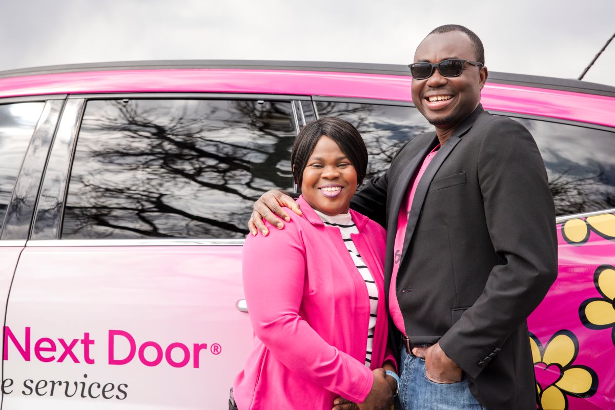 Two Nurse Next Door franchise owners standing beside a pink company car in front of a historic building, symbolizing financial support and success in franchising.