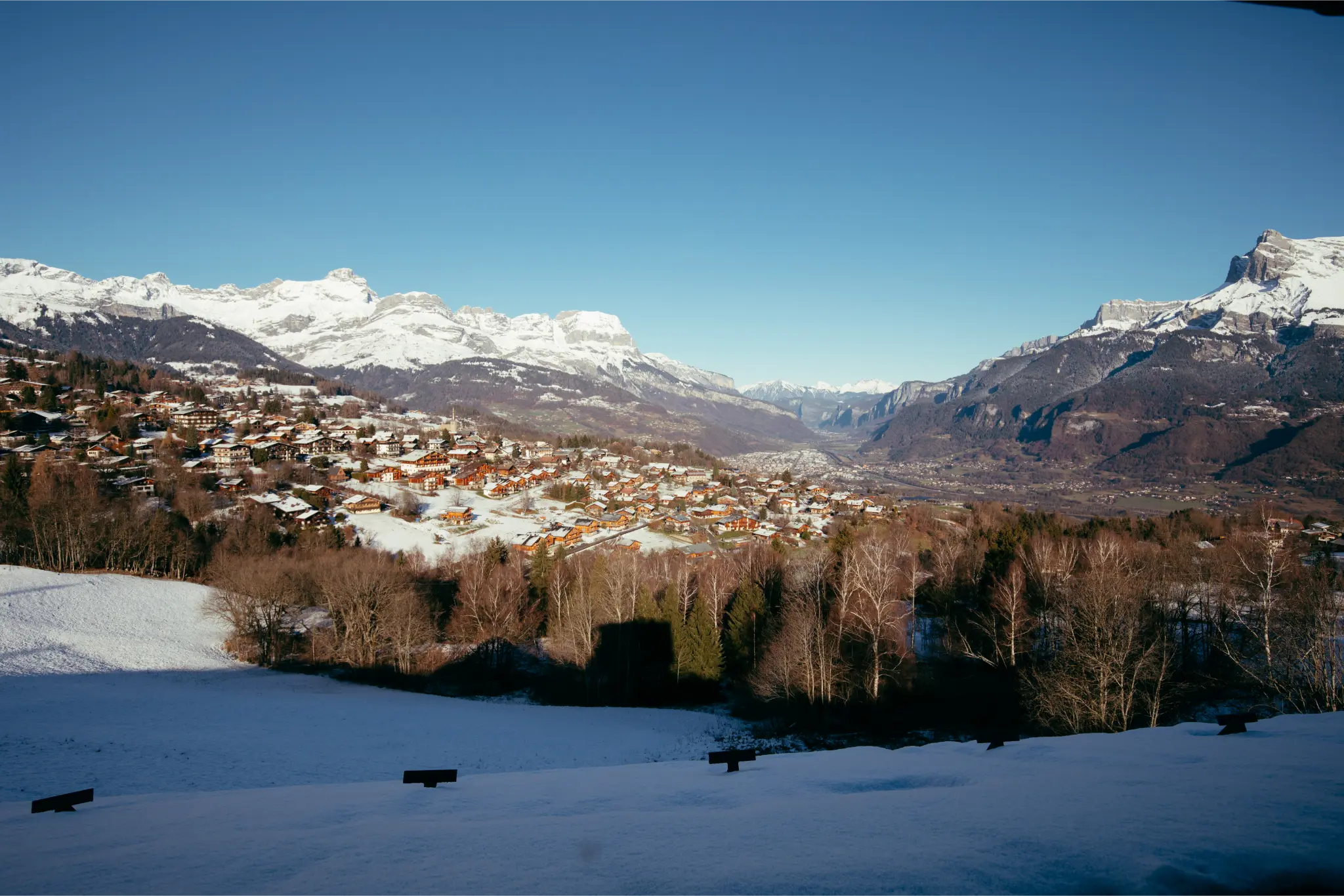 Vue de Combloux sous la neige, avec les toits enneigés et les sommets alpins en arrière-plan.