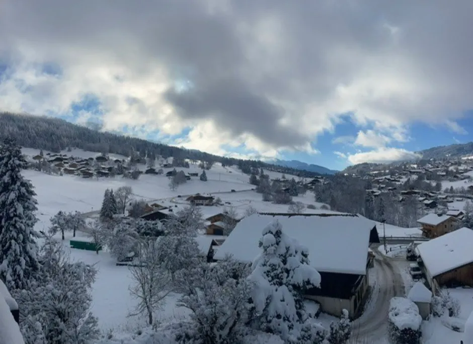 Aerial view of Combloux in the snow, with snow-covered rooftops and Alpine peaks in the background.