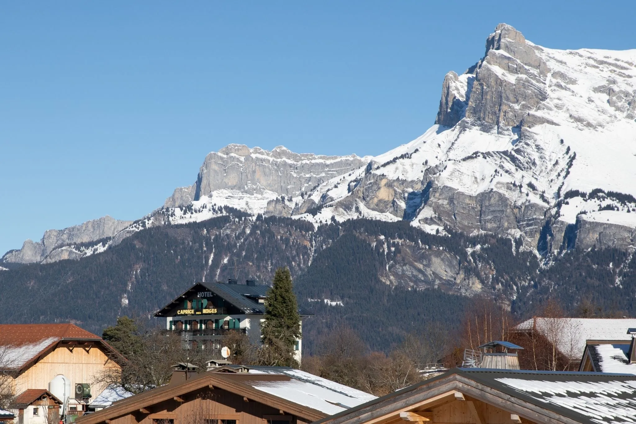 Façade du Caprice des Neiges vue de loin, entourée par les montagnes enneigées de Combloux.