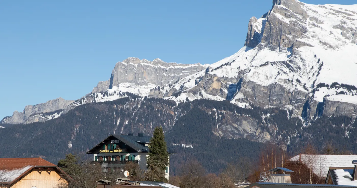 Hotel Le Caprice des Neiges seen from afar, nestled in an alpine landscape with mountains in the background.