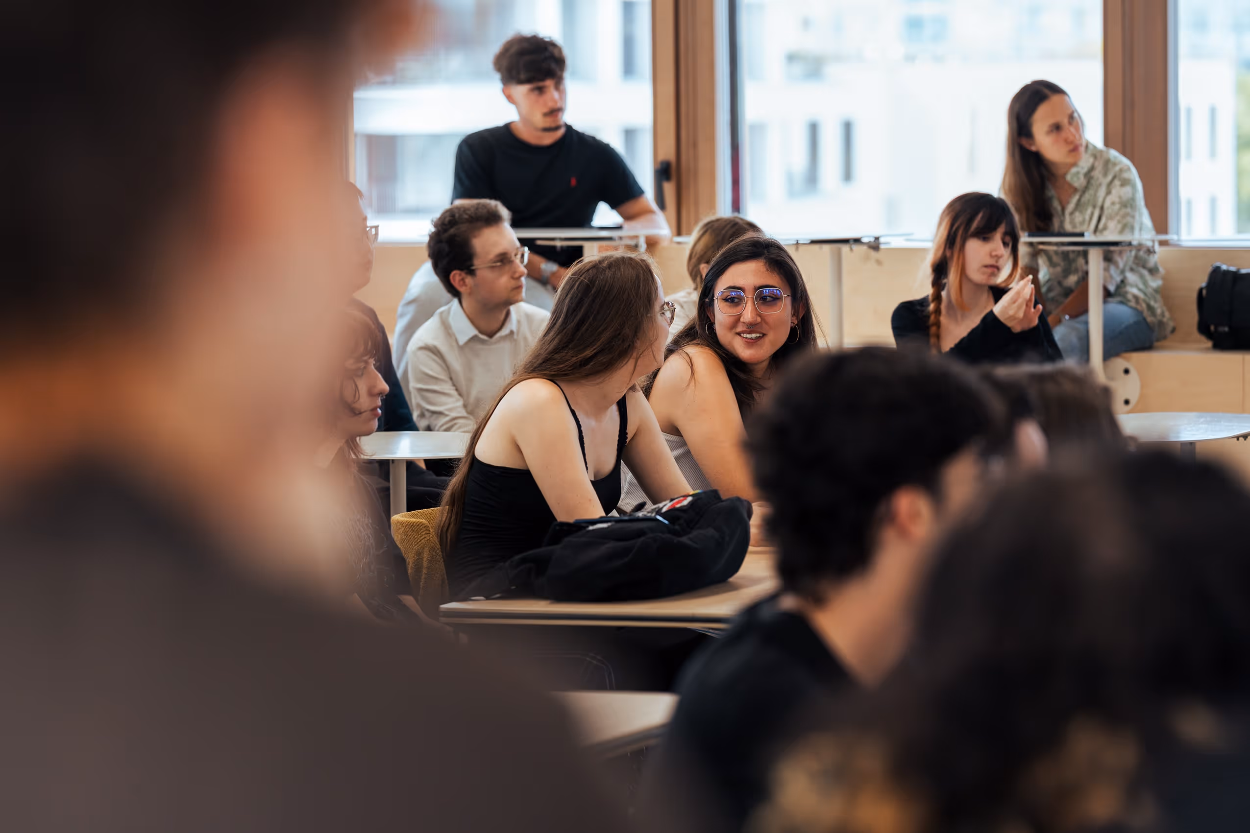 Students are talking and listening in a bright classroom with large windows.