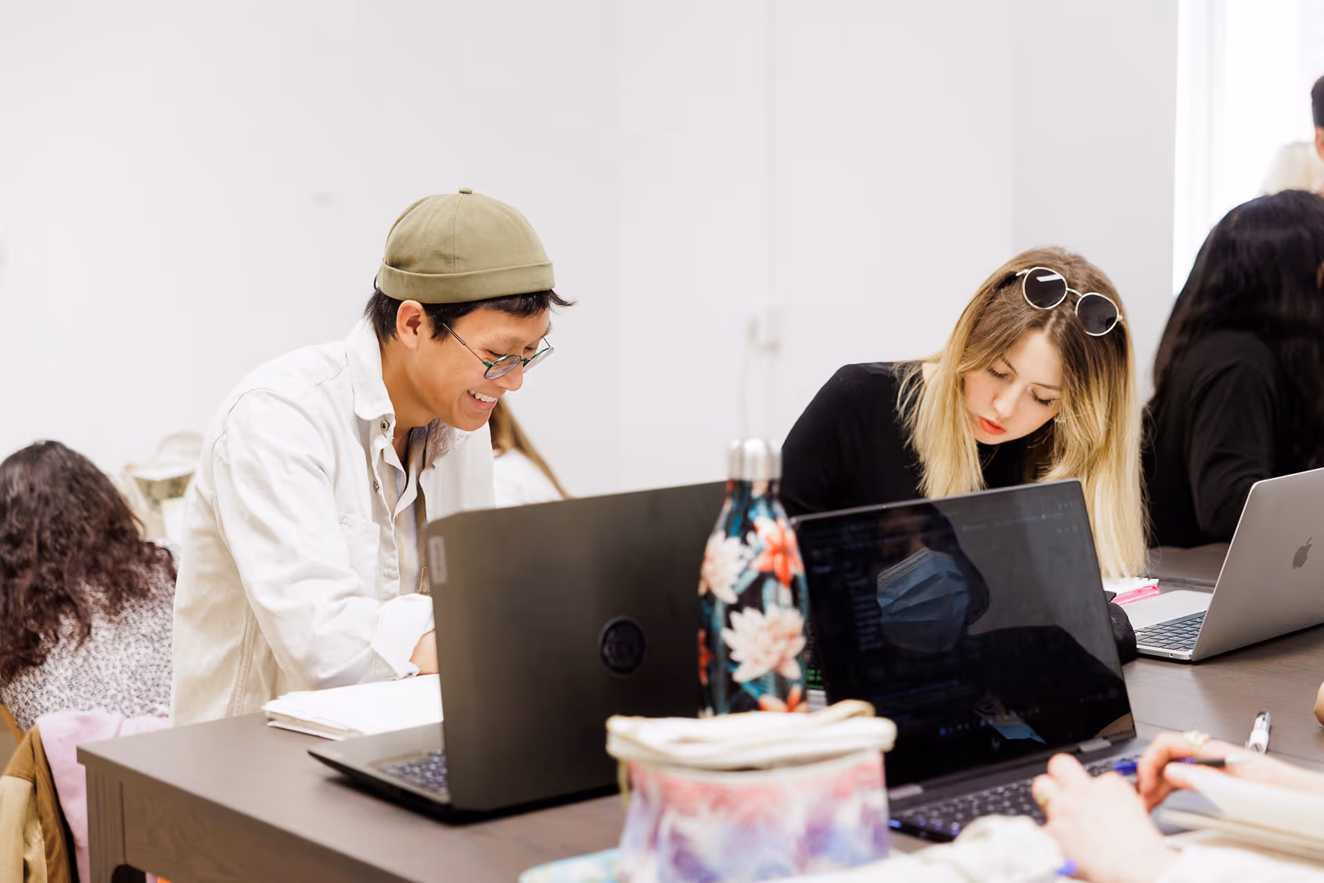 Students work on laptops at a large table in a bright room.