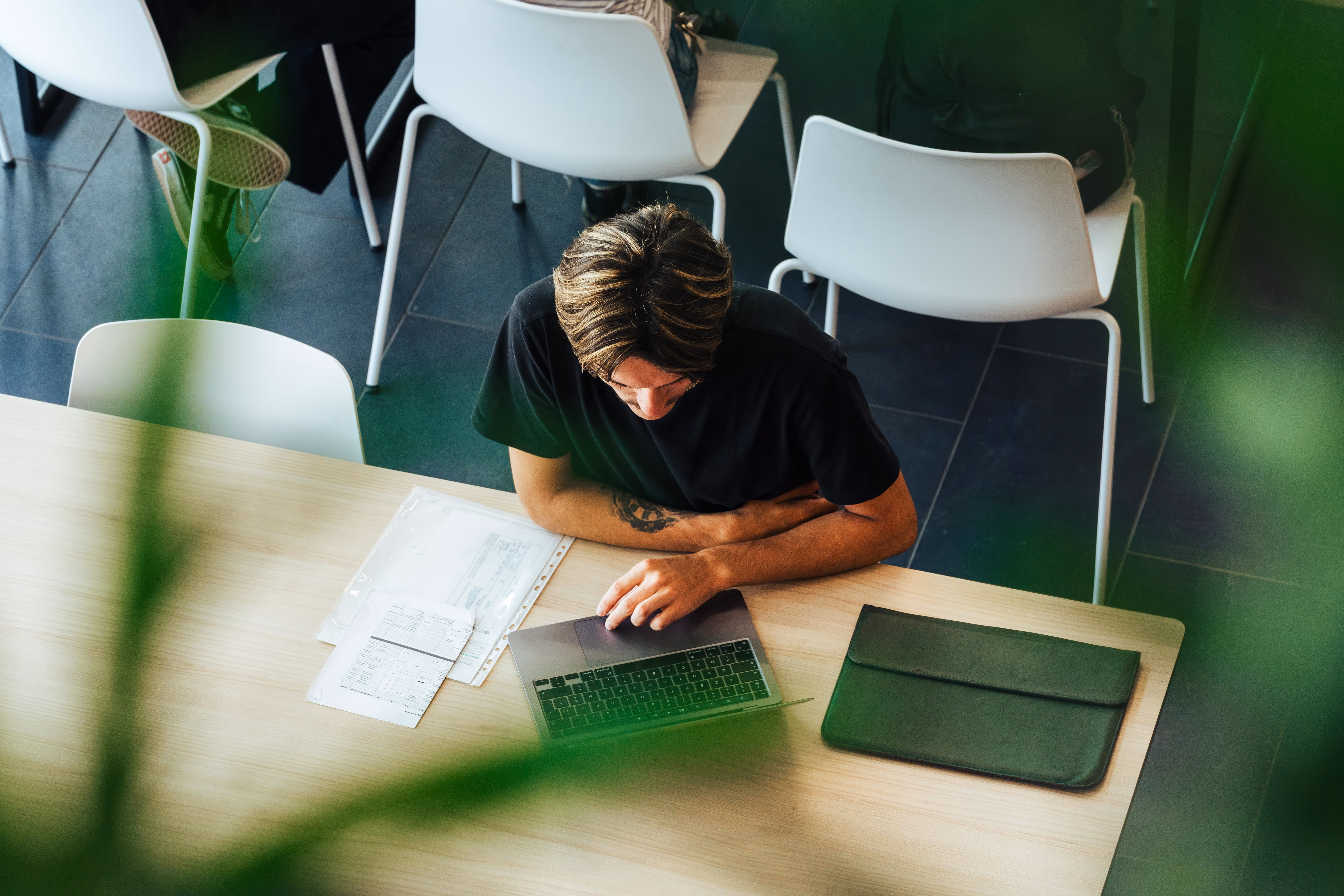Young man sitting at a wooden table working on a laptop computer with documents and a black pouch next to it.