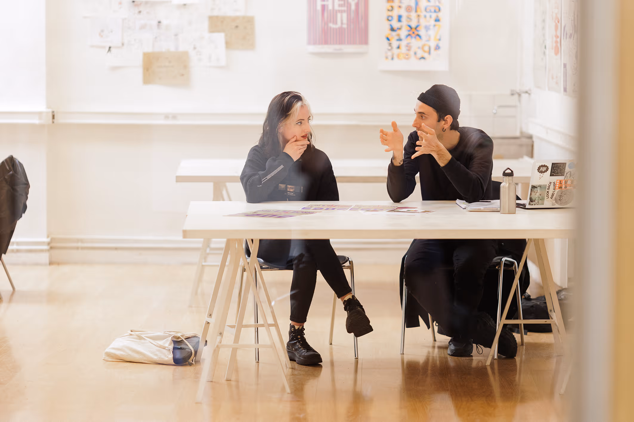 Two people are talking seriously sitting at a white table in a bright room with posters on the walls.