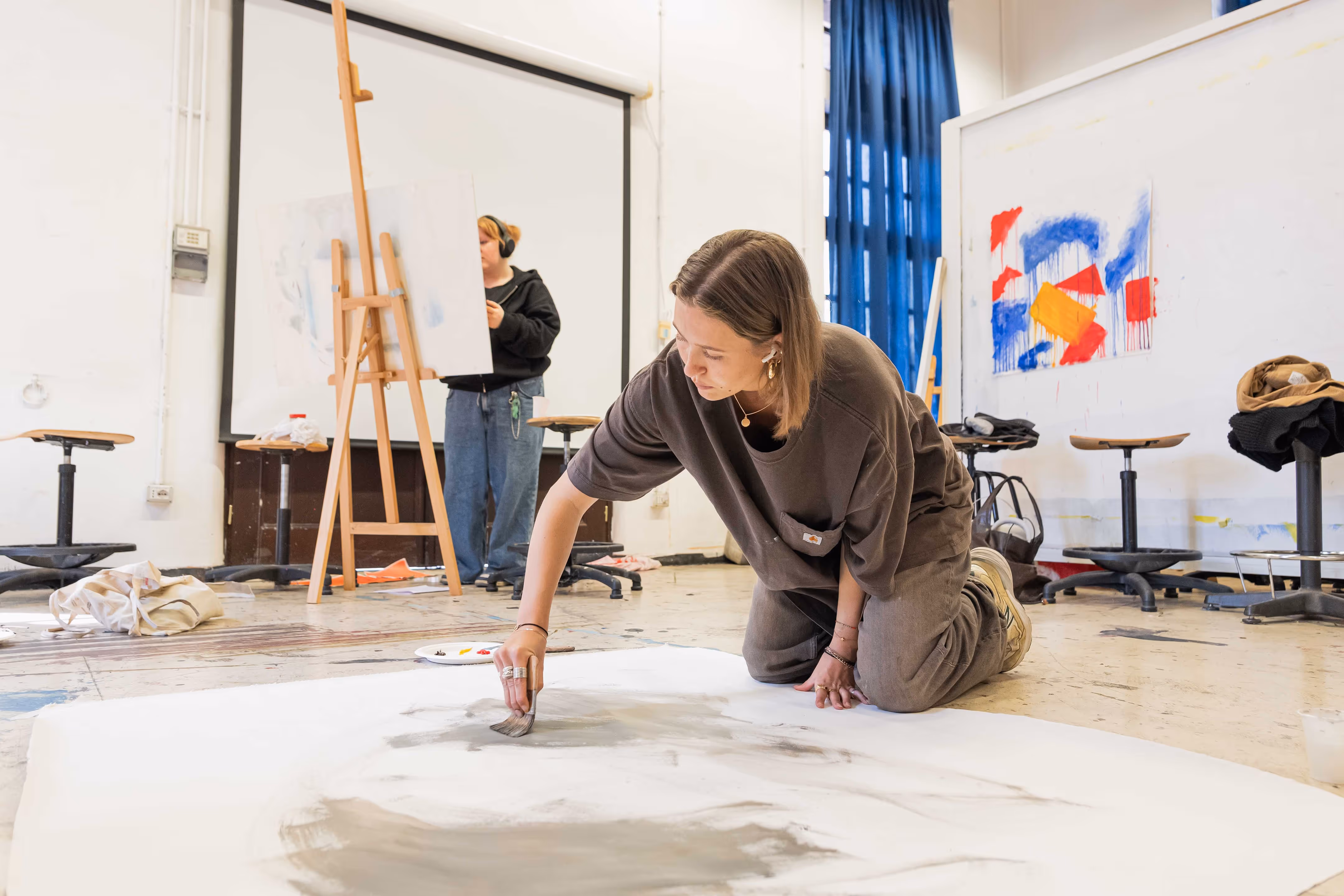 Female student on her knees painted on a large white sheet lying on the ground in an art studio with another student working on an easel in the background.