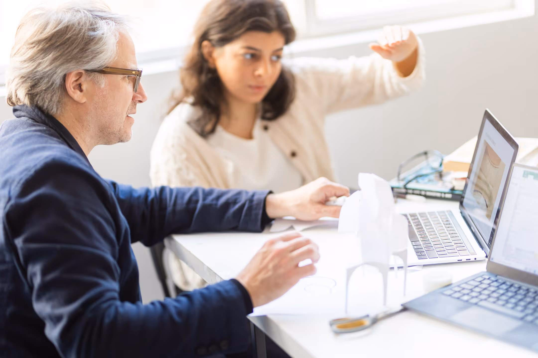 Two people are discussing a project in front of two laptops on a table, with a paper architectural model.