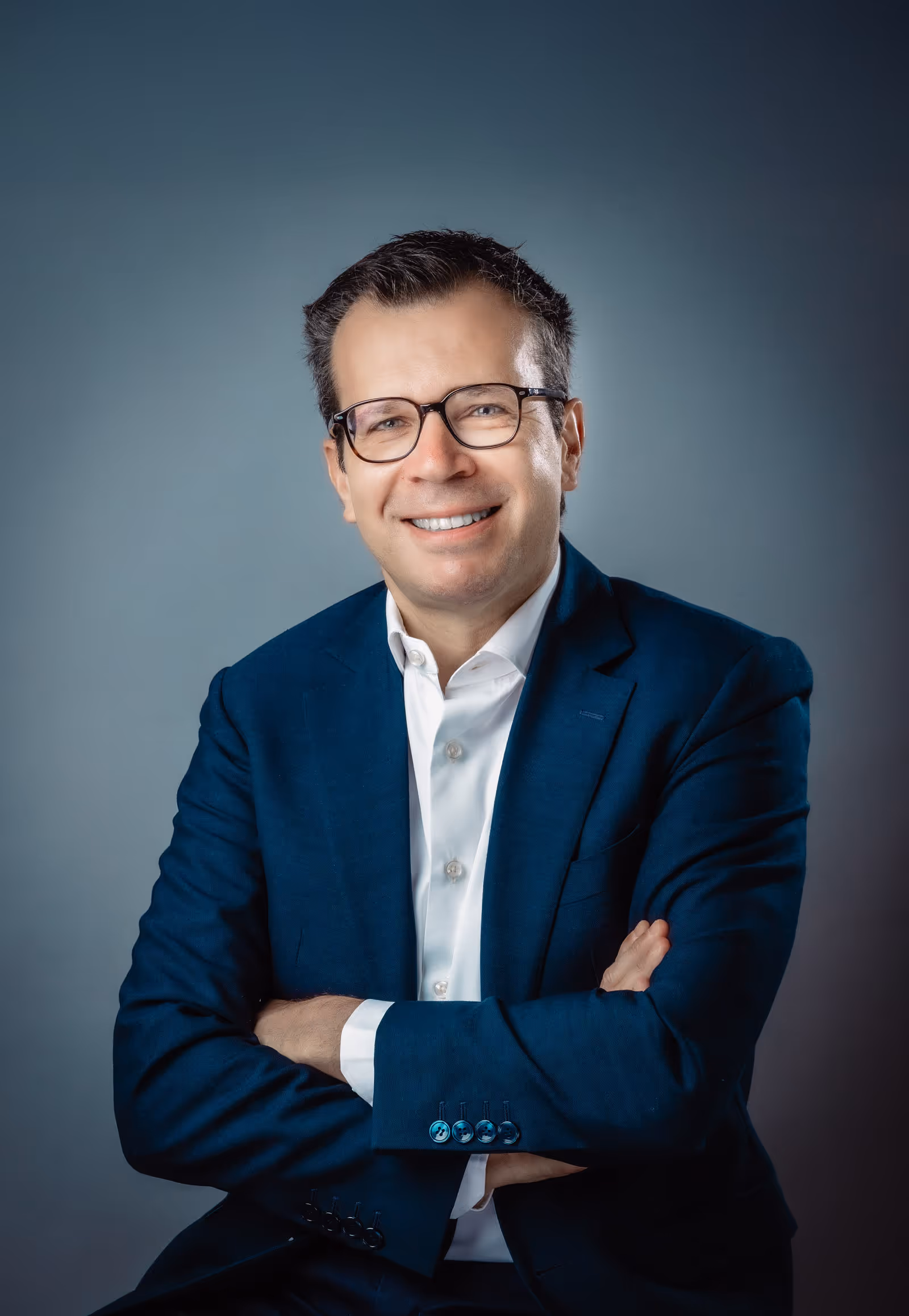 Portrait of Pascal Maury, Group Performance and Human Resources Director, smiling, wearing glasses, a white shirt and a blue suit, his arms crossed in front of a neutral background.