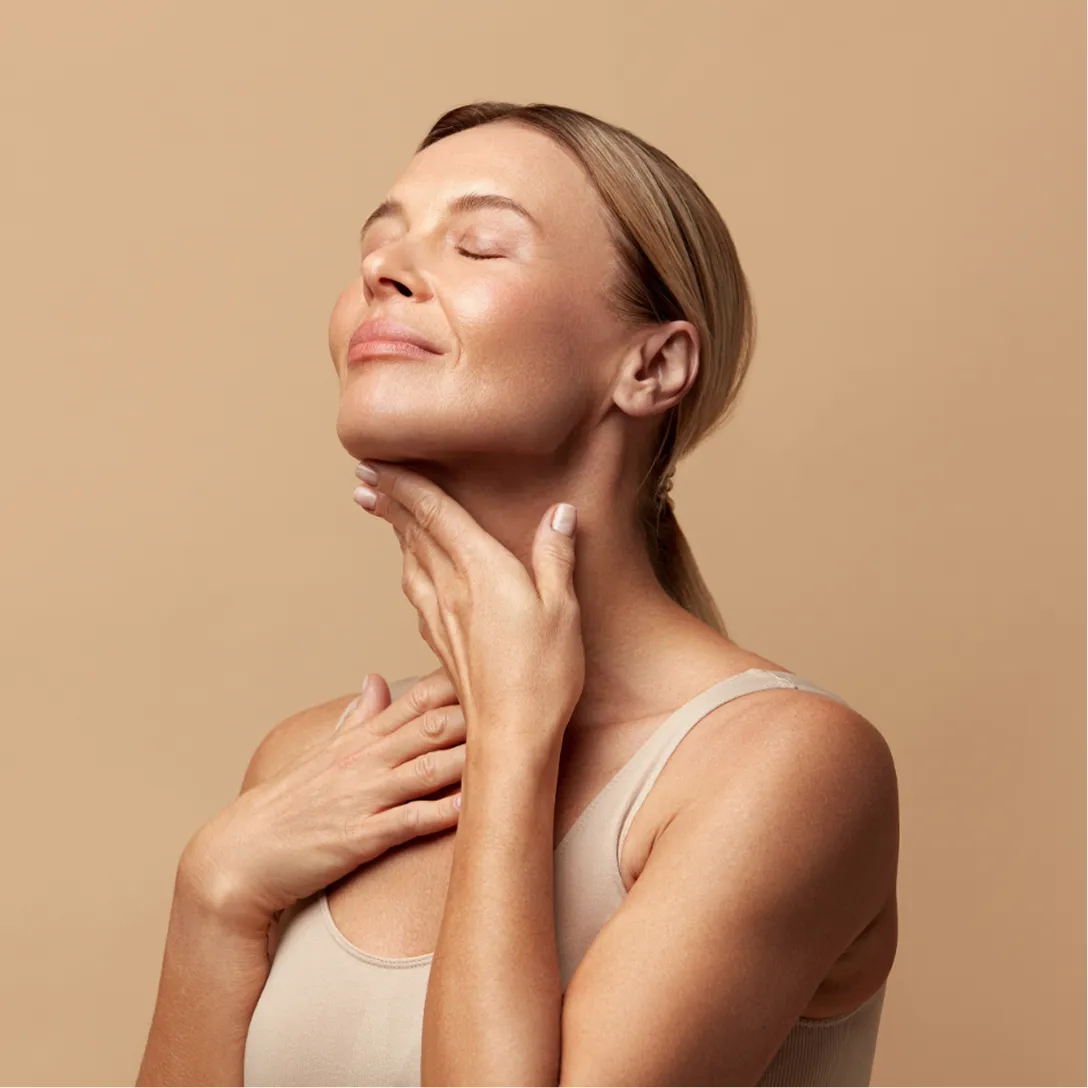 Smiling woman touching her neck with closed eyes against a neutral background.
