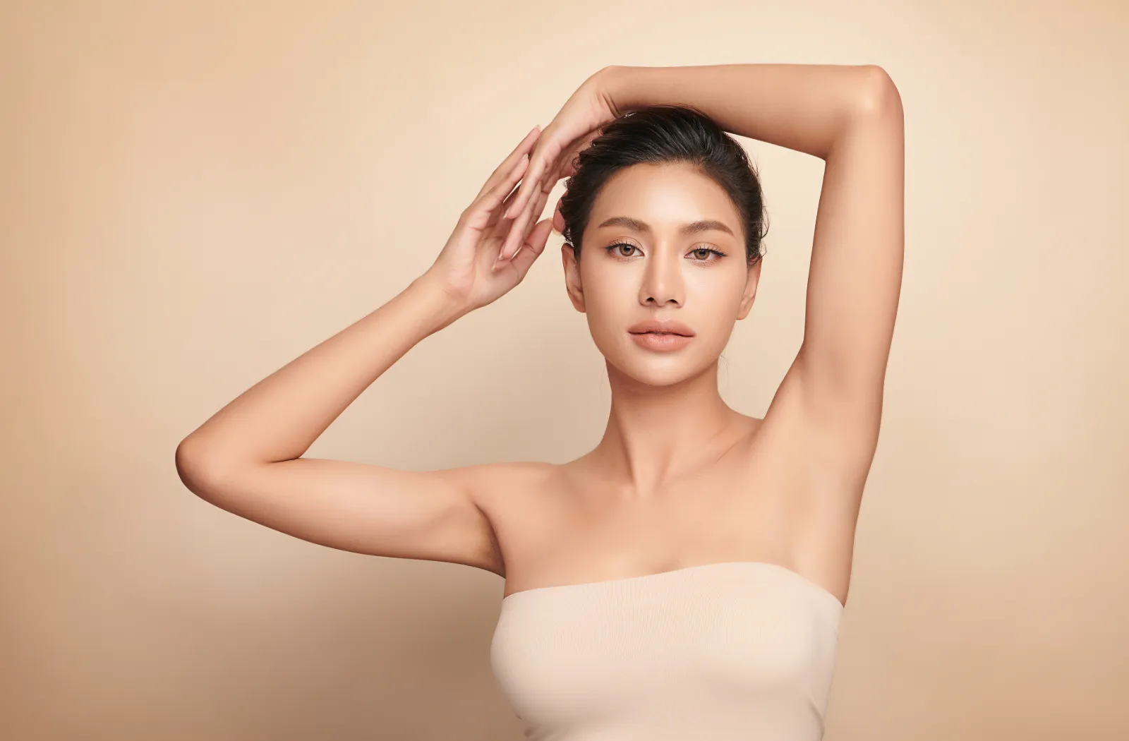 Woman posing gracefully with arms raised, wearing a beige strapless top against a neutral background.