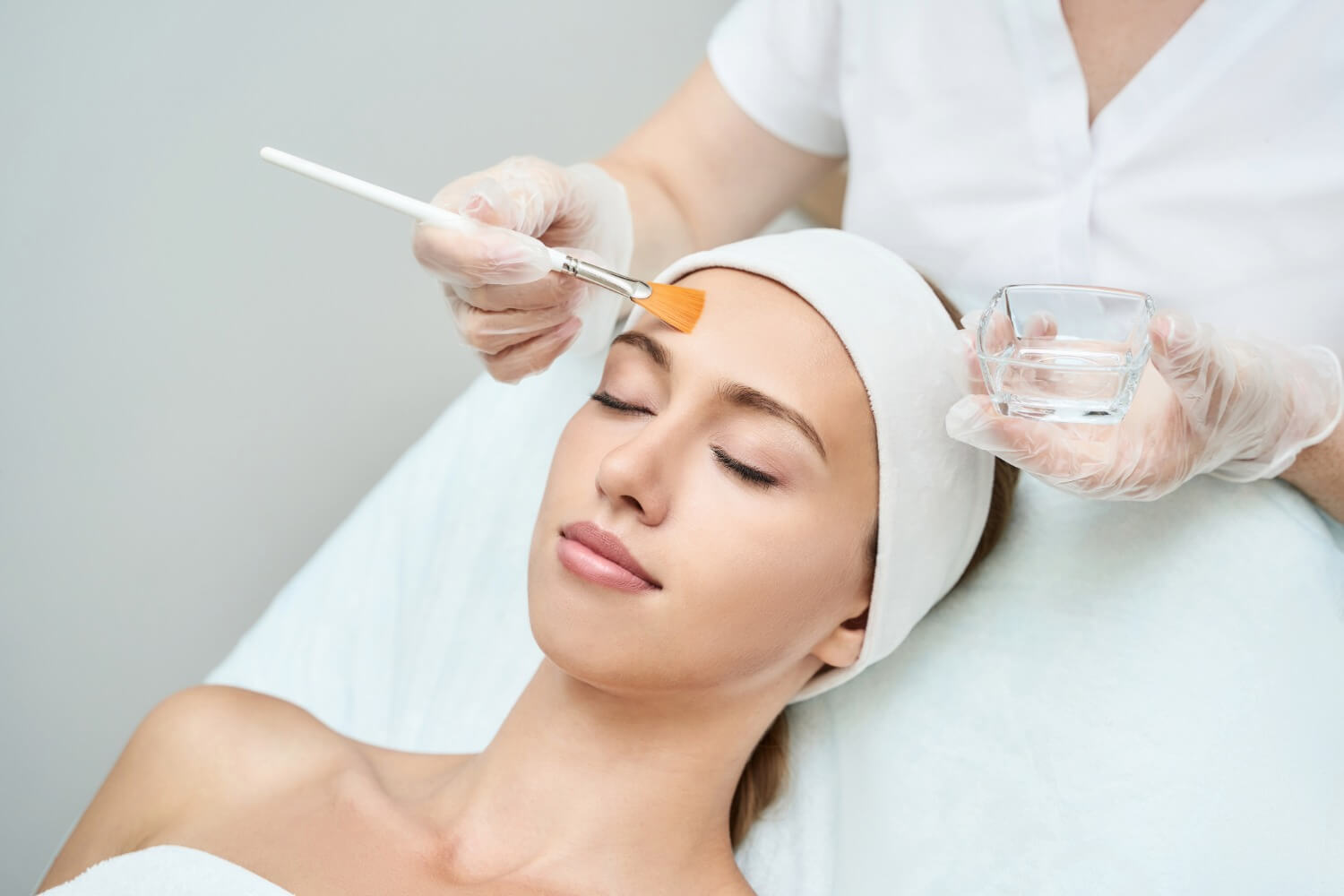 A woman lies with eyes closed while a skincare professional applies a chemical peel to her forehead using a brush; the professional wears gloves and holds a small glass bowl.