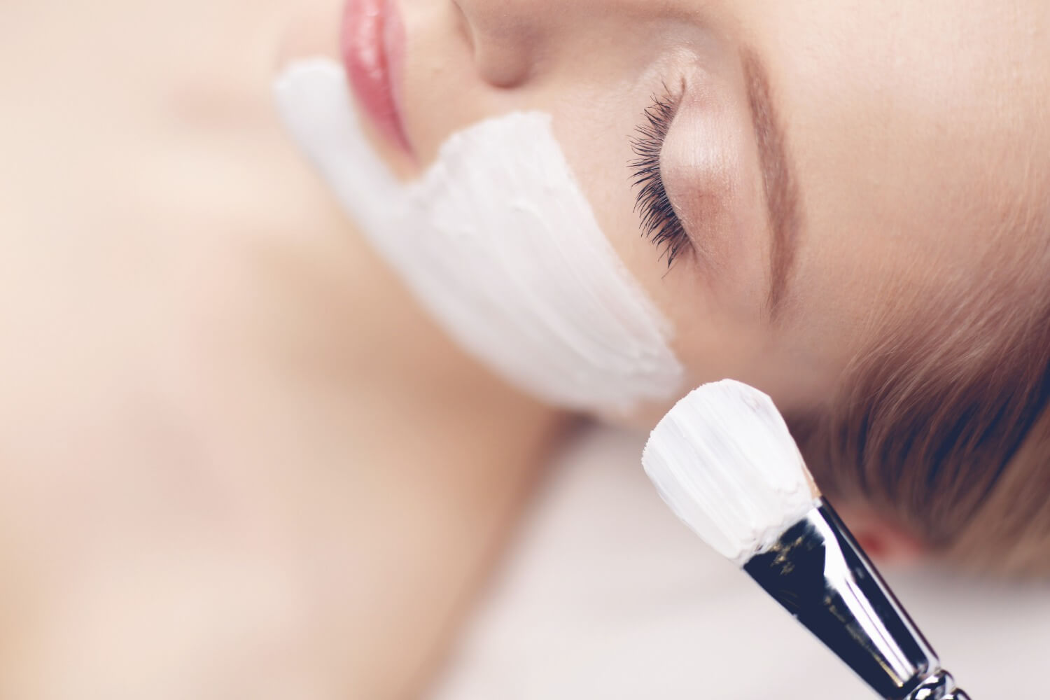 Close-up of a woman with closed eyes lying down as a thick white chemical peel is applied to her cheek with a brush, showing part of the brush in the foreground.