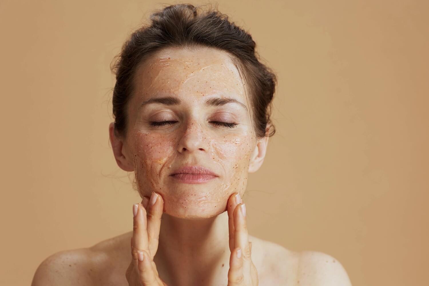 A woman with closed eyes gently massages a translucent exfoliating chemical peel with fine particles onto her face, set against a neutral beige background.