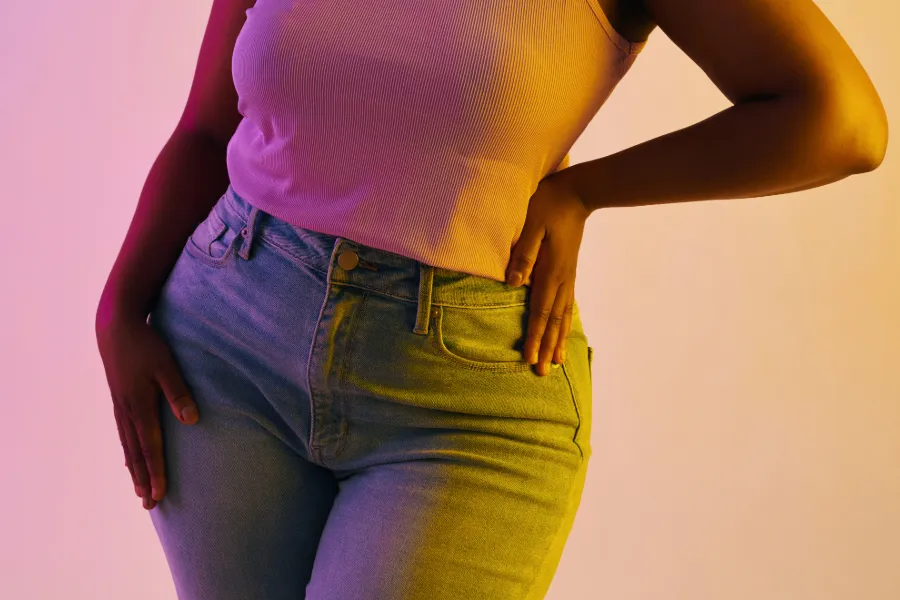 Person wearing high-waisted jeans and a sleeveless top with one hand on hip and the other resting on thigh, illuminated by colorful pink and yellow lighting.