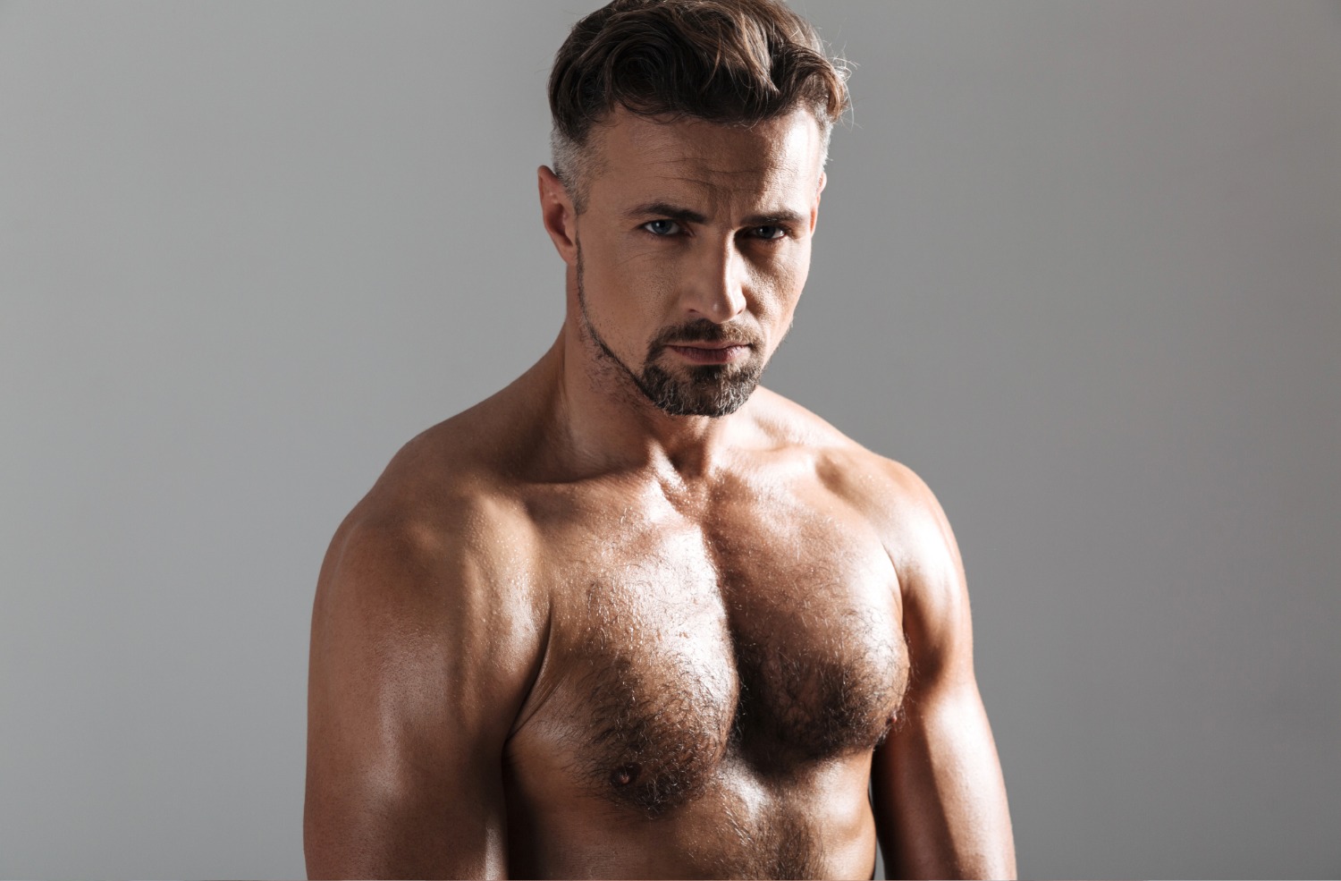 Muscular shirtless man with short styled hair and a trimmed beard against a plain gray background.