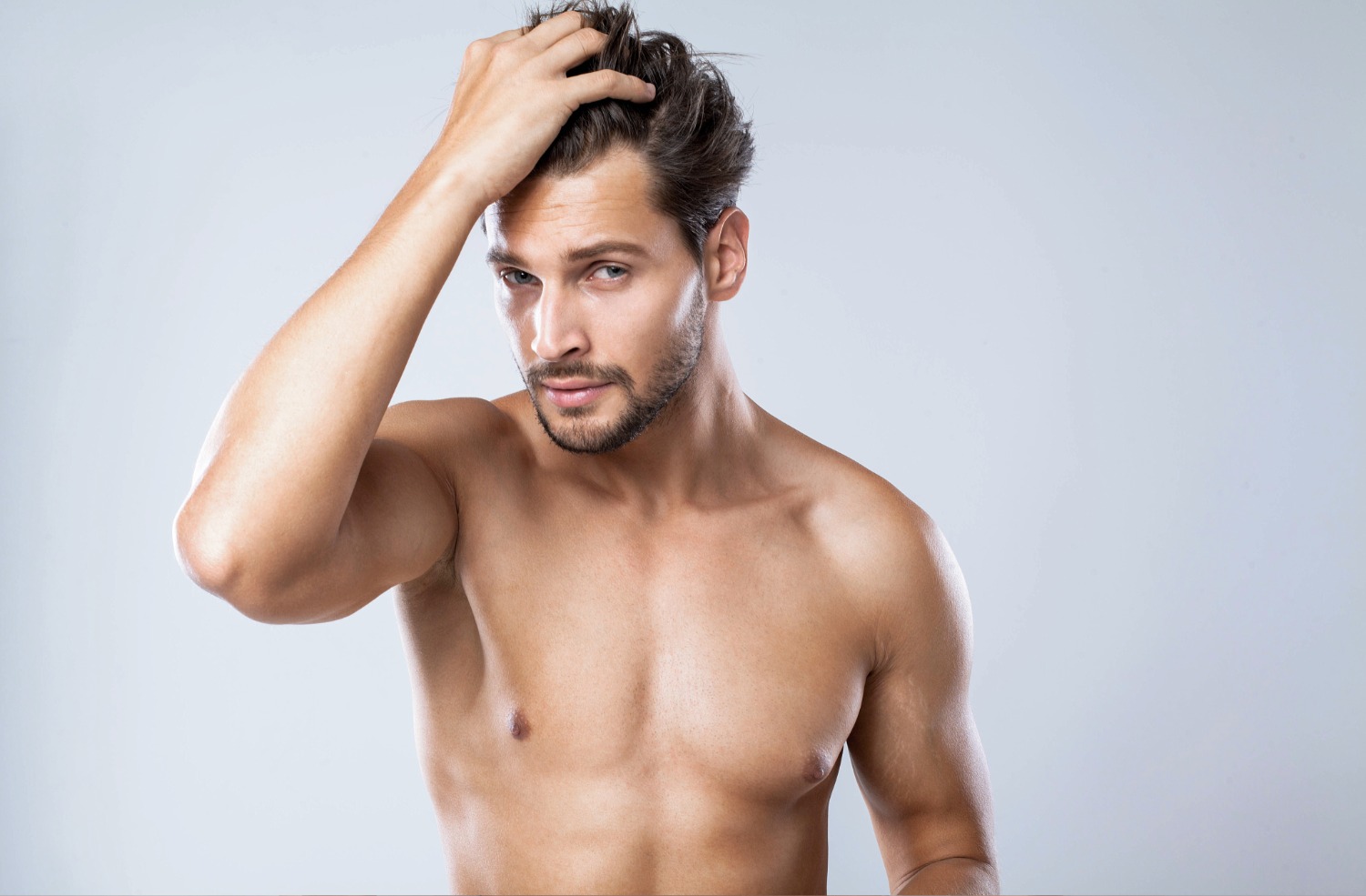 Shirtless man with light beard running hand through his dark hair against a plain light gray background.
