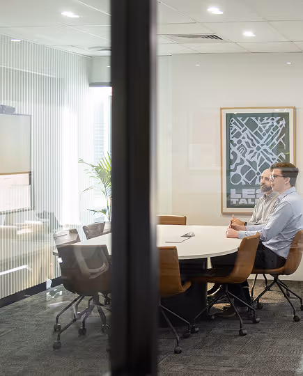 View through a glass meeting room showing a team member working solo at a table in a calm, focused studio environment—capturing clarity and intention in the workspace.