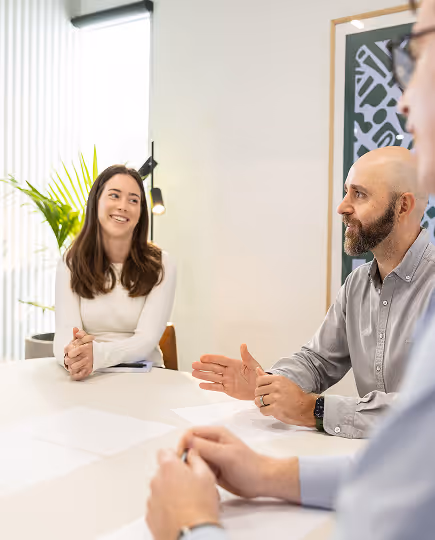 Team gathered in a roundtable discussion, sharing ideas in a bright and relaxed office space with plants and clean design—reflecting open communication and shared thinking.
