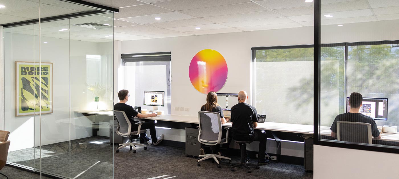 Modern design studio with a collaborative team working at desktop stations in a bright, glass-partitioned office space, featuring contemporary wall art and natural lighting—reflecting a focused and creative agency environment.