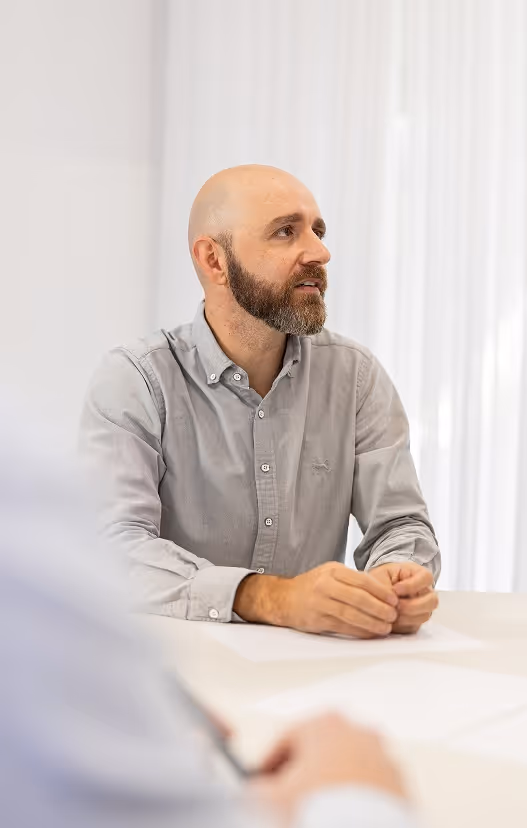 Professional in a button-up shirt seated at a white table in a well-lit meeting room, actively engaged in a conversation, symbolising strategic thinking, leadership, and a human-centred approach to collaboration.