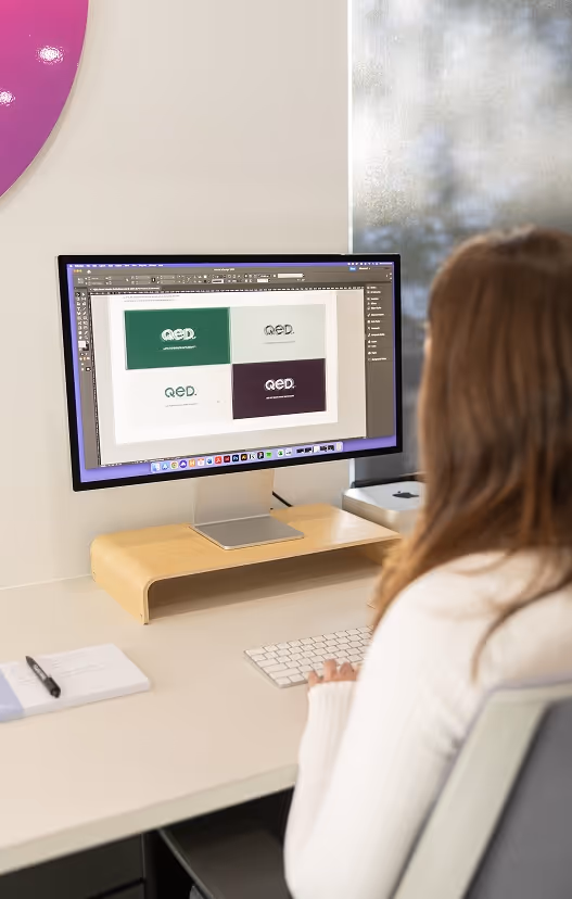 Designer working on a brand identity layout using Adobe InDesign, showcasing logo variations and colour blocks for a corporate brand on a high-resolution screen, reflecting strategic branding work in progress.