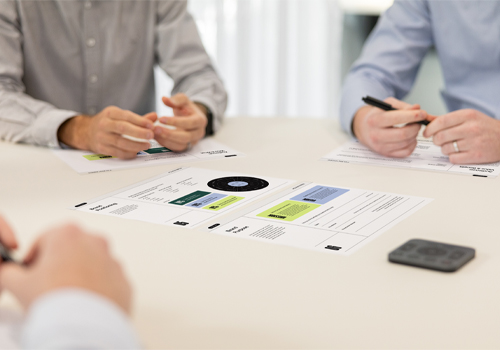 Three people seated at a table reviewing printed charts and documents with pens in hand.