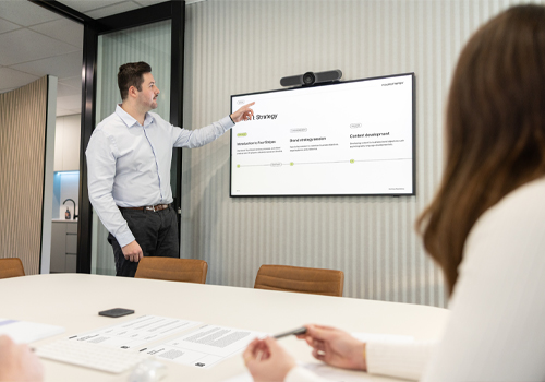Man standing and pointing at a wall-mounted screen displaying a digital strategy presentation while two people watch from a conference table.