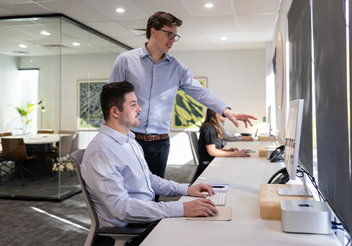 Two men in an office setting, one seated at a computer and the other standing and pointing at the monitor.
