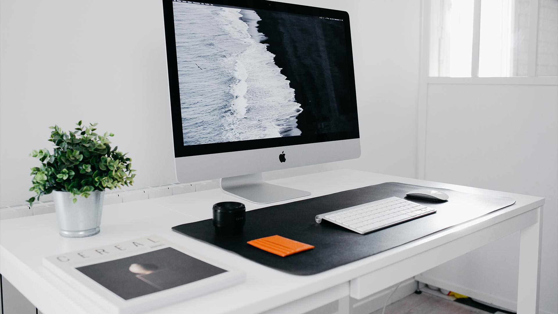 Apple iMac, keyboard, computer mouse, camera objective, flowerpot and Cereal magazine on the white table.