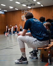 Im Vordergrund ein Basketballspieler auf der Bank und im Hintergrund Spieler auf dem Platz.
