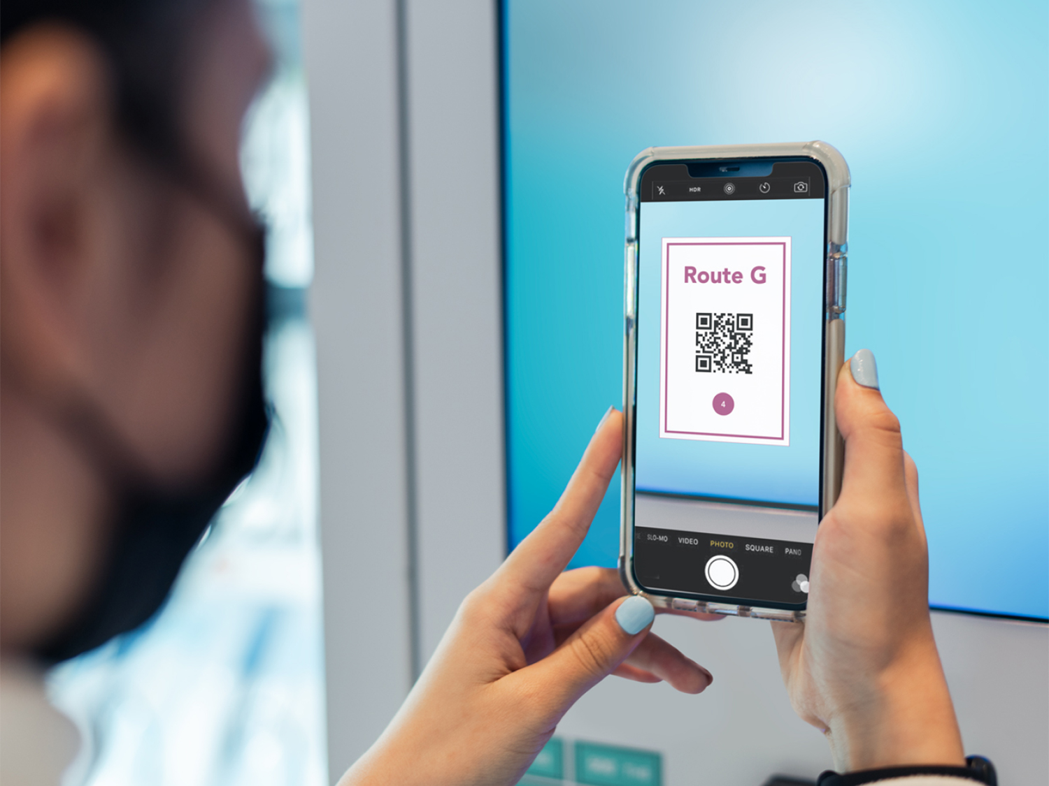 A person wearing a mask and holding up their phone to scan a QR code from the wall