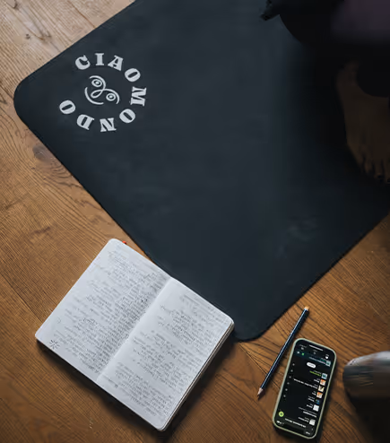 Tappetino yoga nero con logo "Ciao Mondo", affiancato da un quaderno aperto pieno di appunti, una penna e uno smartphone su un pavimento in legno.