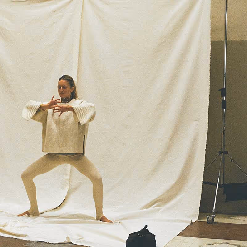 Donna in posa di movimento consapevole o danza meditativa, su sfondo neutro con telo bianco e attrezzatura da studio fotografico.