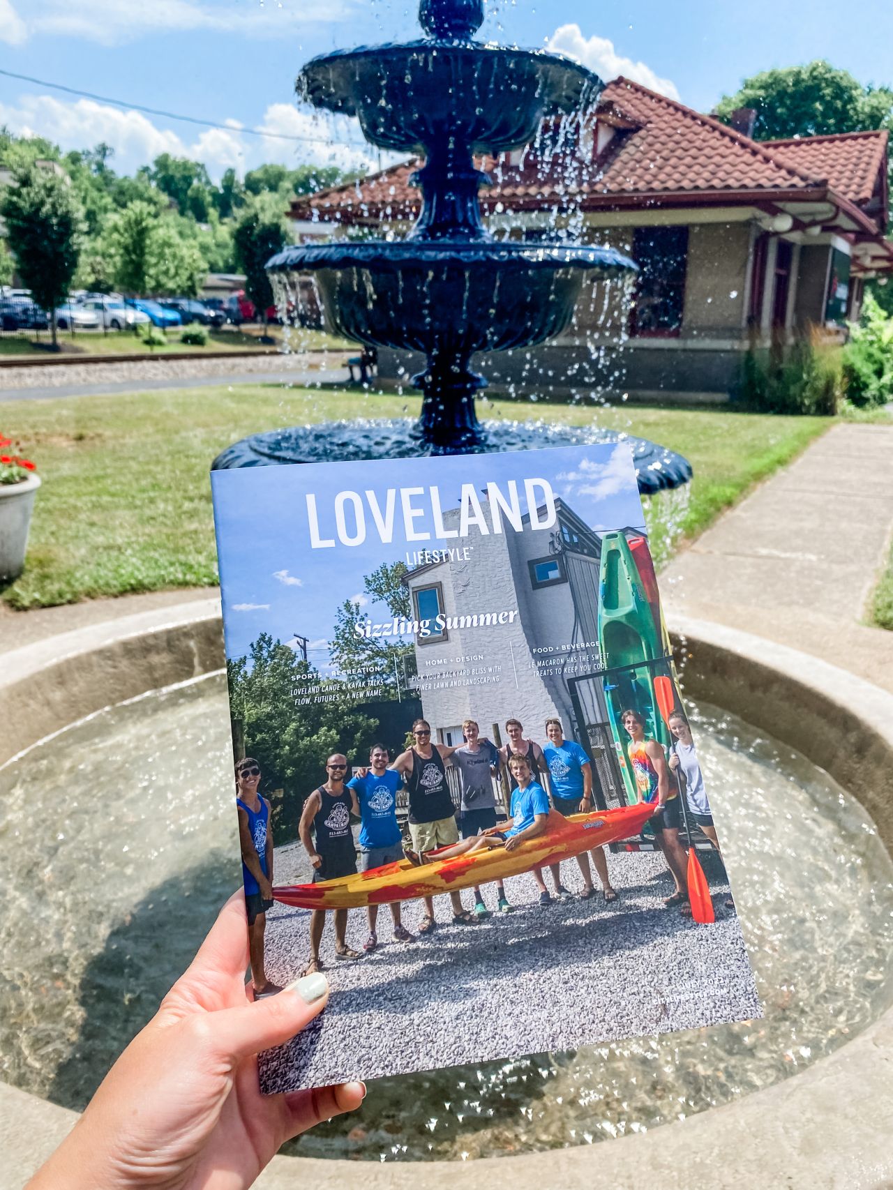 A hand holds up a Loveland City Lifestyle magazine in front of a bubbling tiered fountain. The magazine cover features a group of smiling people standing outdoors, holding a kayak, with the headline “Sizzling Summer.” In the background, a sunny courtyard with trees, a sidewalk, and a red-roofed building can be seen.