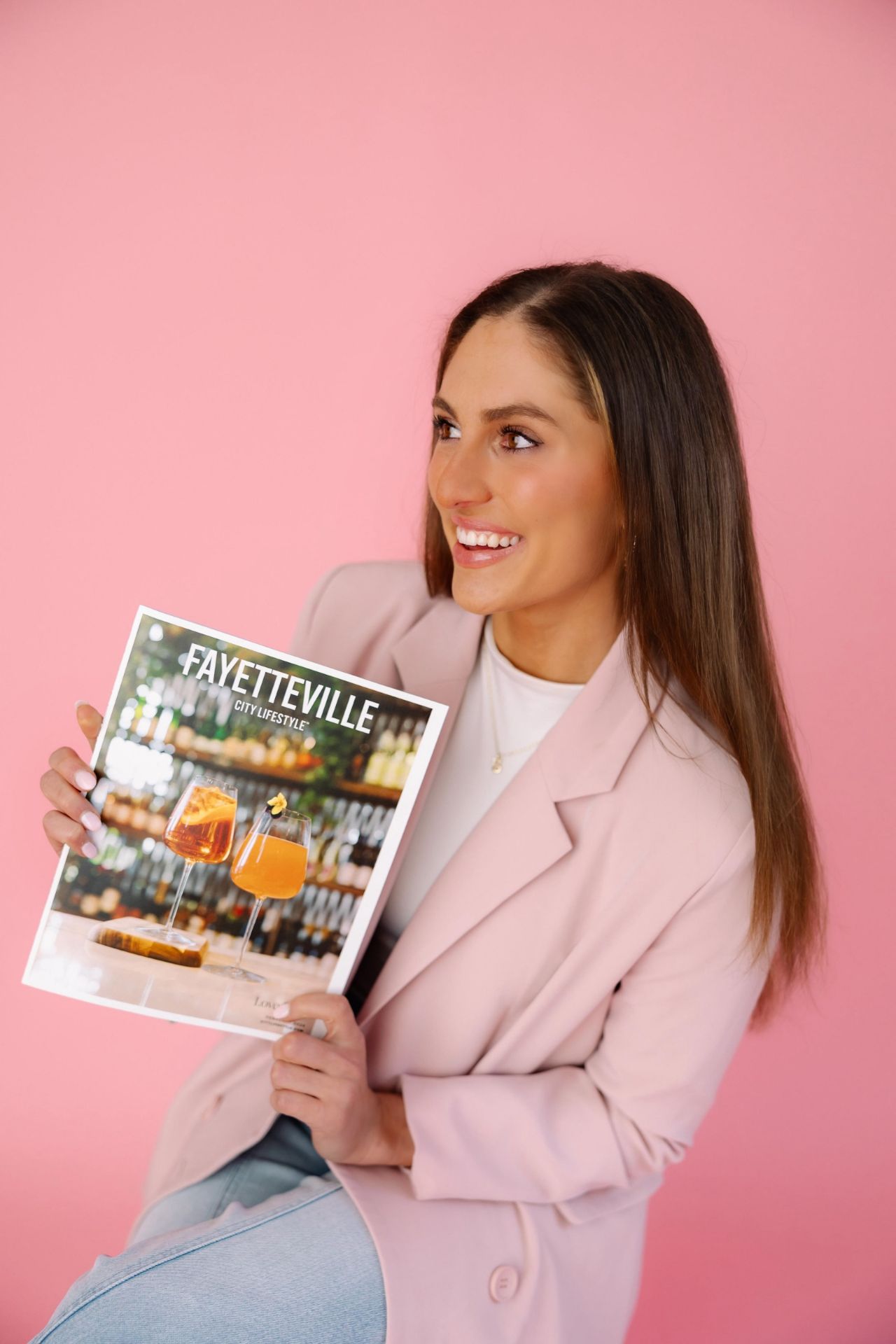 A smiling woman in a pale pink blazer holds up a Fayetteville City Lifestyle magazine against a pink background. The magazine cover features two orange cocktails and a wooden snack board in front of a blurred bar shelf display.