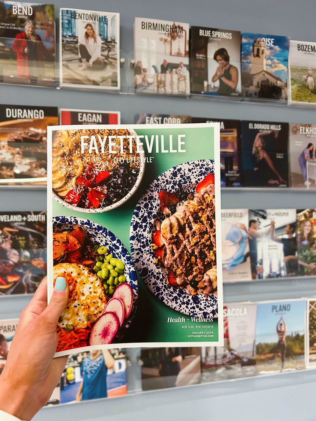 A hand with light blue nail polish holds up an issue of Fayetteville Lifestyle magazine in front of a display wall filled with other city editions. The cover features vibrant, colorful bowls of healthy food against a green background, highlighting the theme "Health + Wellness."