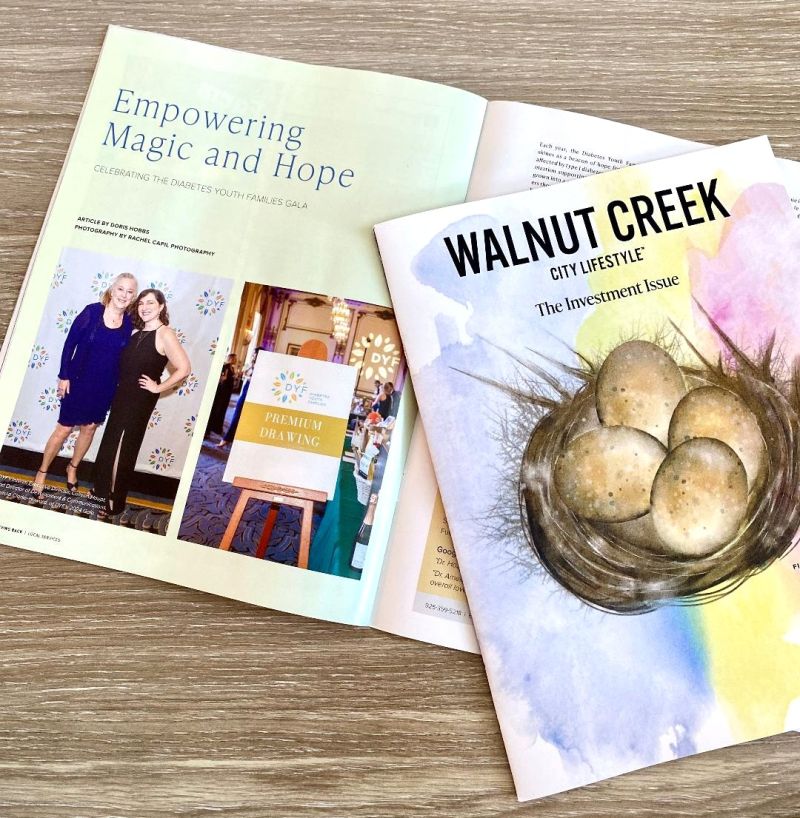 A copy of Walnut Creek City Lifestyle magazine lies atop an open issue. The cover, labeled “The Investment Issue,” features a painted bird’s nest cradling golden eggs against a soft watercolor background. The open spread highlights a feature titled “Empowering Magic and Hope” with event photos from a youth gala. The scene is set on a light wood surface, evoking a calm and hopeful tone.