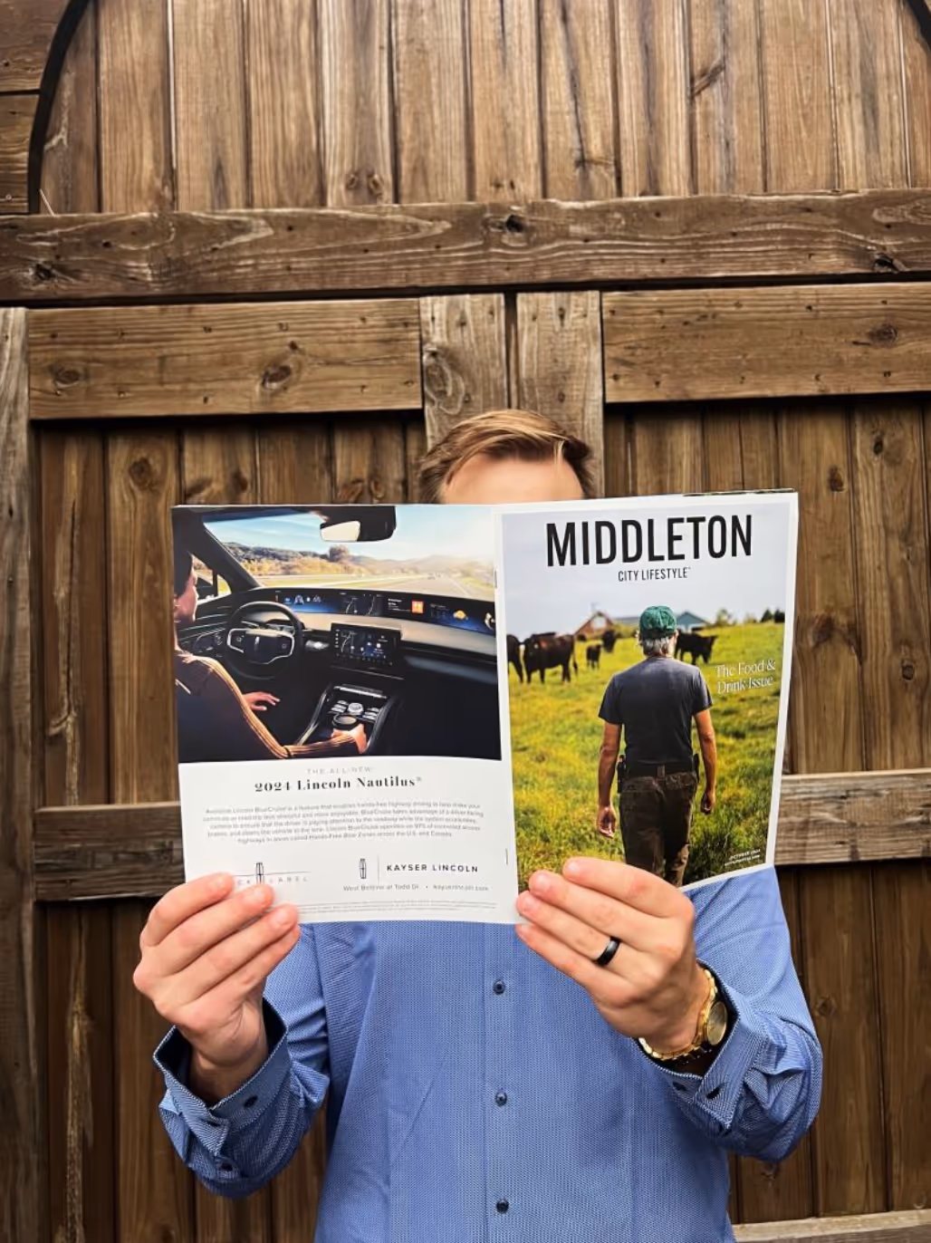 A man in a blue shirt stands in front of a rustic wooden wall, holding up an open Middleton City Lifestyle magazine that covers most of his face. The front cover shows a man walking through a grassy field with cows, under the theme “The Food & Drink Issue.” The back cover features an ad for the 2024 Lincoln Nautilus.