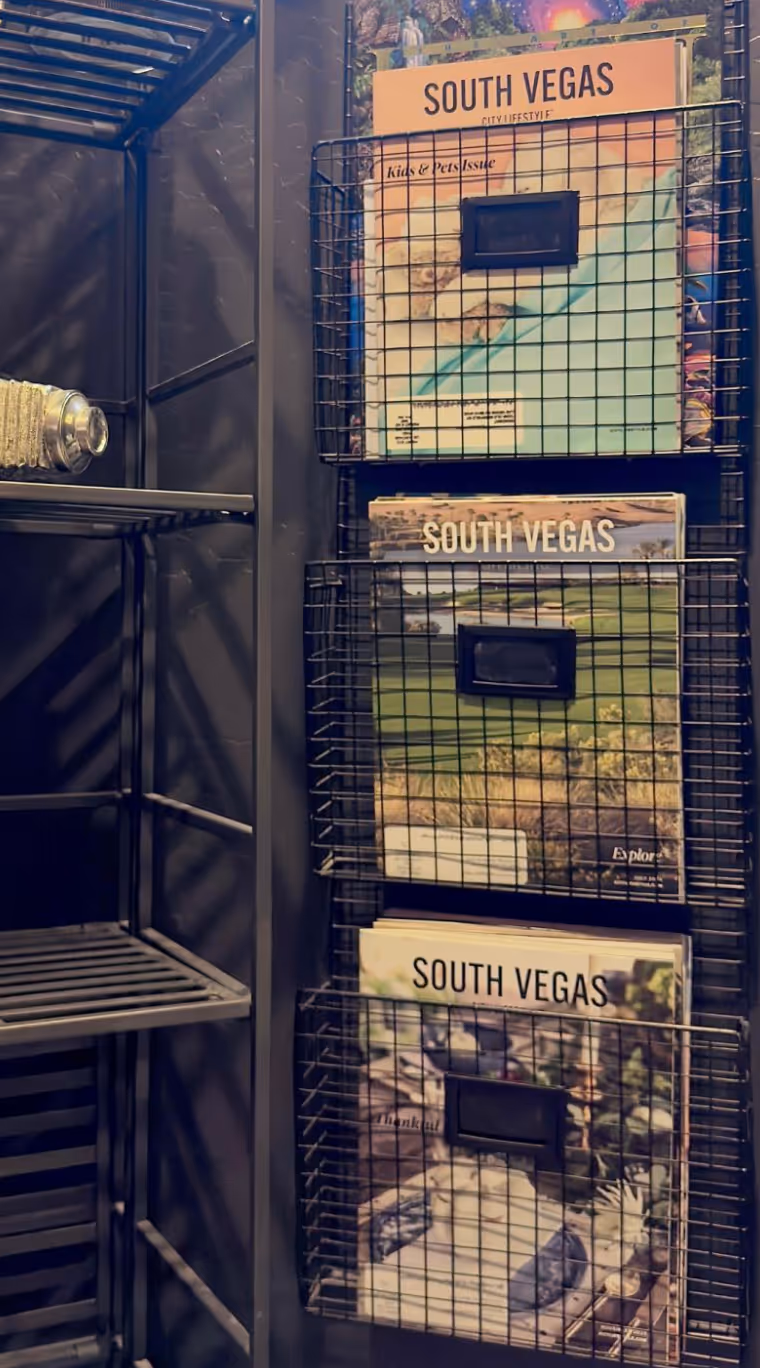 A set of South Vegas City Lifestyle magazines are neatly displayed in black wire wall racks against a dark shelving unit. The magazine covers feature a vibrant green golf course landscape under a clear sky. Nearby, metal shelves and a glass water bottle add an industrial touch to the scene, suggesting a stylish waiting area or retail space.
