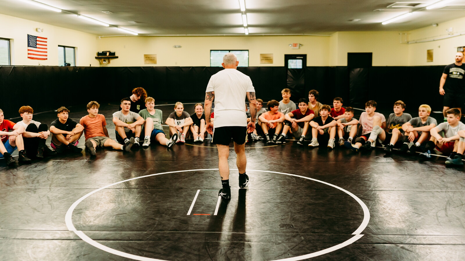 A wrestling coach stands center mat with his back to the camera, addressing a group of young athletes seated around the circle in a brightly lit gym. The room exudes focus and tradition, capturing a moment of mentorship and intensity at The House of Krause in Parkland, Missouri.