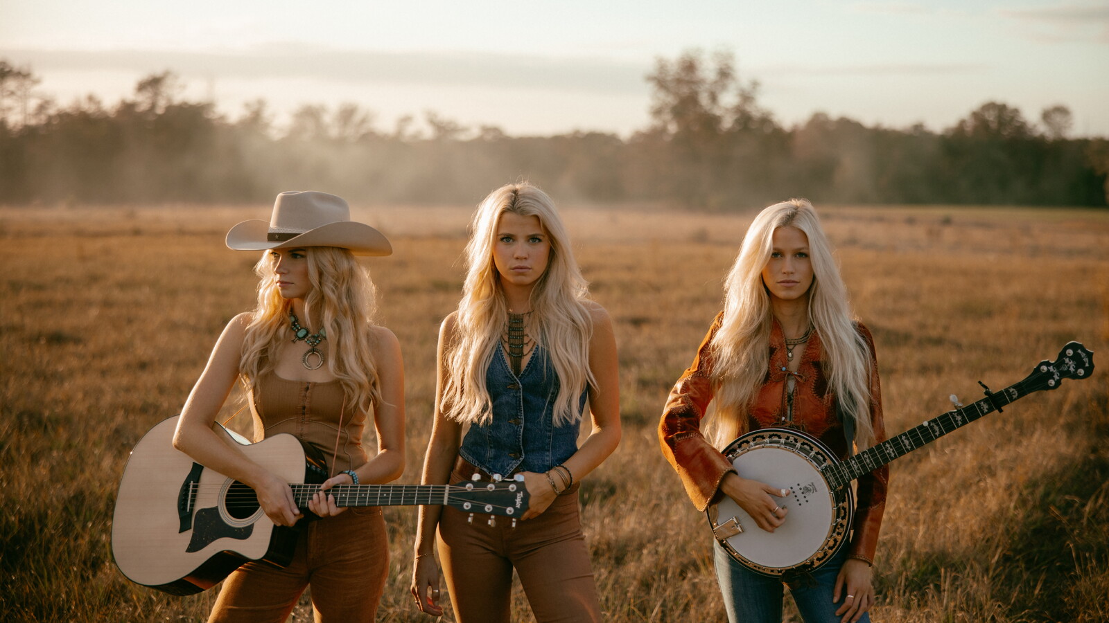 Three blonde women standing in a golden field at sunset, dressed in western-style outfits. Two hold instruments — a guitar and a banjo — while the center woman stands with hands at her sides, exuding confidence. A nod to country music and Southern storytelling.