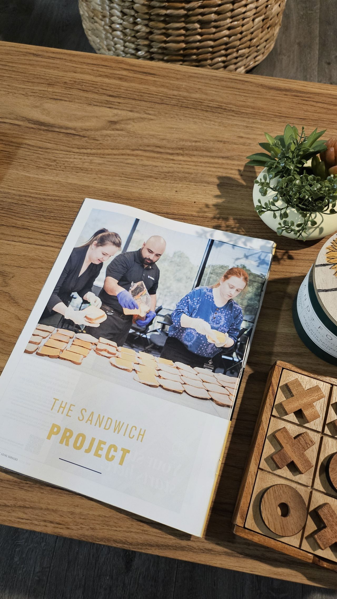 An open City Lifestyle magazine lies on a wooden table next to a potted plant and a wooden tic-tac-toe game. The featured article, titled “The Sandwich Project,” shows three volunteers assembling sandwiches in an assembly line with stacks of bread spread across the table. Natural light from large windows brightens the scene, capturing a moment of community service and collaboration.