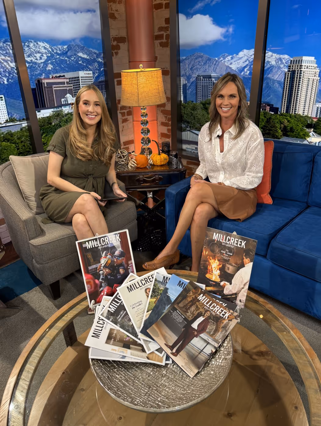 Two women sit on a talk show set with a view of a city skyline and mountains in the background. A table in front of them displays several Millcreek City Lifestyle magazines featuring various covers, including a hockey player, a fireplace scene, and professional portraits. The atmosphere is warm and inviting, with autumn decor and a cozy lamp between the chairs.
