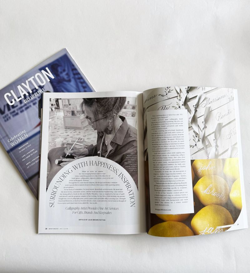 An issue of Clayton City Lifestyle lies on a white surface, opened to a feature spread. The left page shows a black-and-white image of a woman drawing or writing, with the headline "Surrounding with Happiness + Inspiration" below her. The right page includes two columns of text alongside a collage featuring handwritten script and bright yellow lemons labeled with words like “love,” “harmony,” and “joy.” The closed cover underneath showcases the magazine’s title in bold white on blue, with the tagline “Celebrating Women.”
