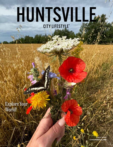 Cover of the July 2025 issue of Huntsville City Lifestyle. A hand holds a vibrant wildflower bouquet — including red poppies, daisies, and Queen Anne’s lace — with a black-and-white butterfly resting on the flowers. In the background, a golden wheat field stretches toward a line of green trees under a blue sky. Text on the cover reads: “Explore Your World!”