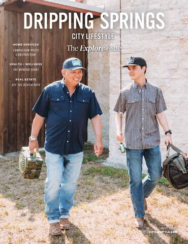 Cover of Dripping Springs City Lifestyle, July 2025 issue, titled “The Explore Issue.” It features two men walking side by side outside in casual clothing and baseball caps, carrying fishing or outdoor gear. One is older, wearing a navy shirt and jeans, and the other is younger, in a short-sleeve plaid shirt and jeans. They appear to be heading out for an outdoor adventure, walking past a stone wall and wooden door.