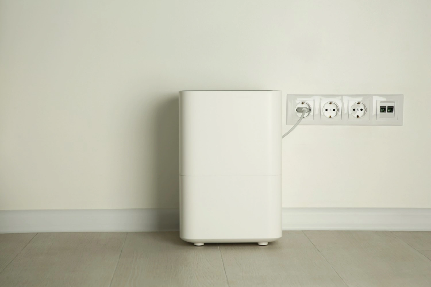 A simple, modern, white humidifier plugged into a wall outlet in a room with a light wood floor and white walls.